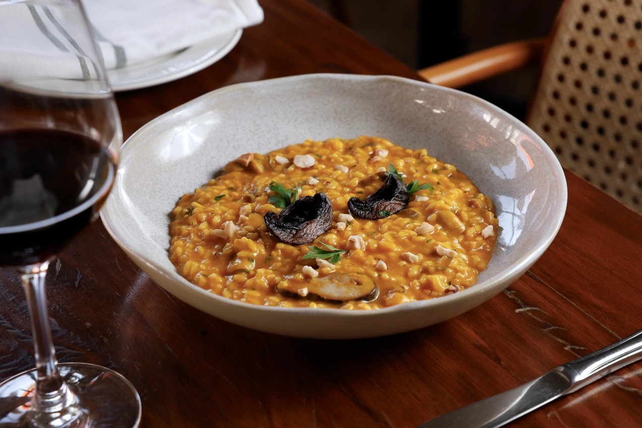 Risoto amarelo com cogumelos, castanhas e taça de vinho sobre mesa de restaurante.