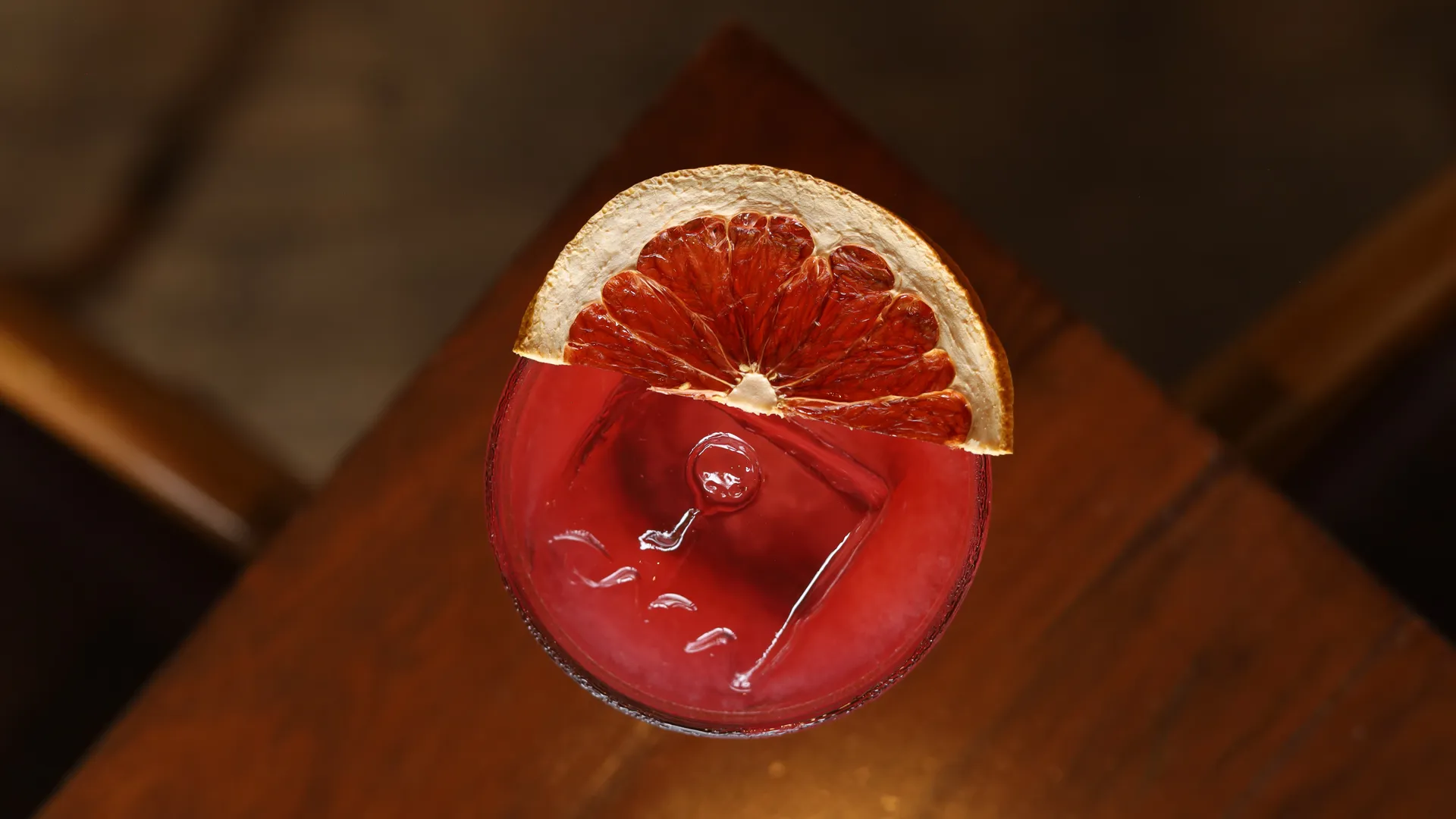 Drink vermelho com fatia de laranja desidratada sobre mesa de madeira.