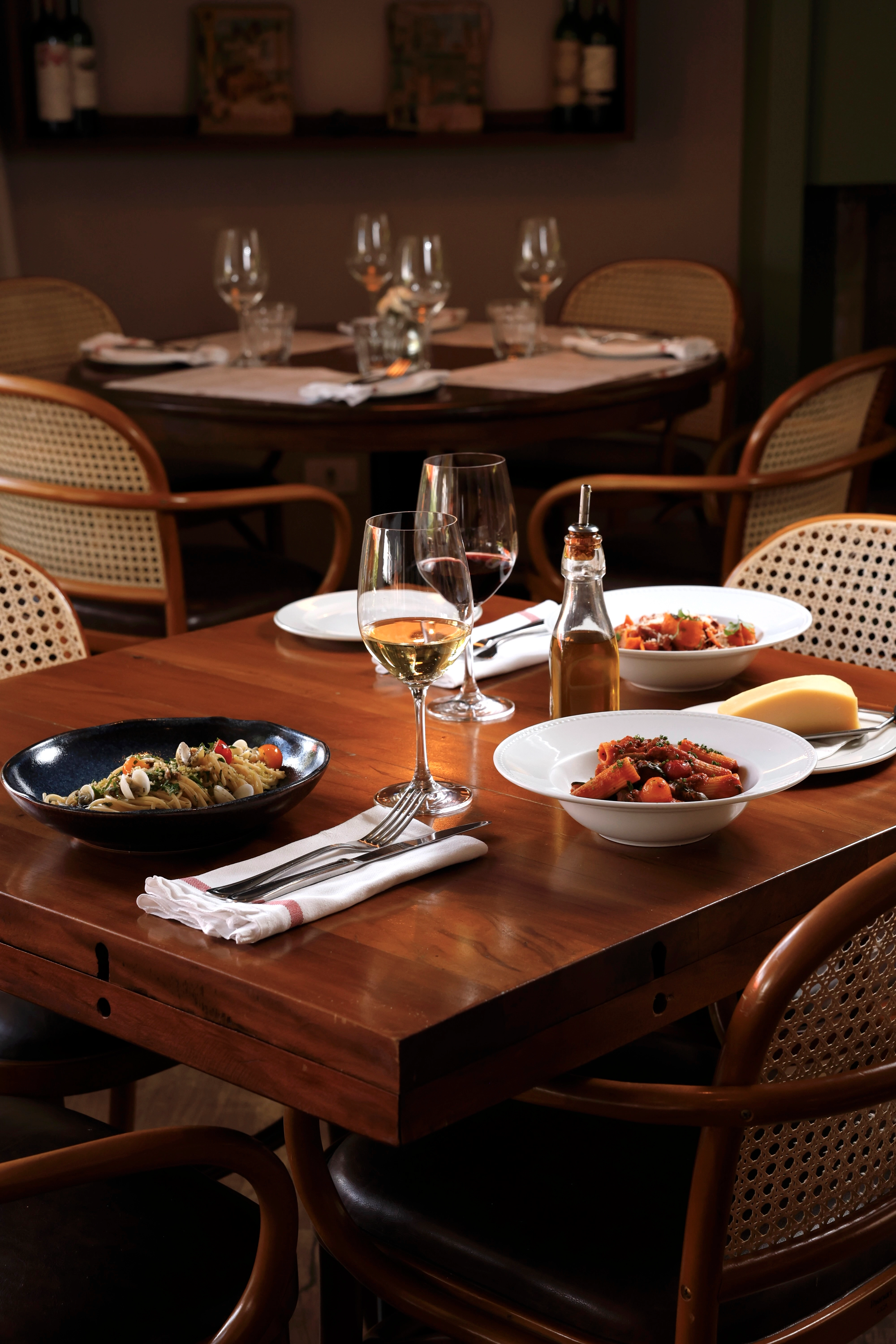 Mesa de restaurante com massas, vinho e lugares preparados para receber clientes.