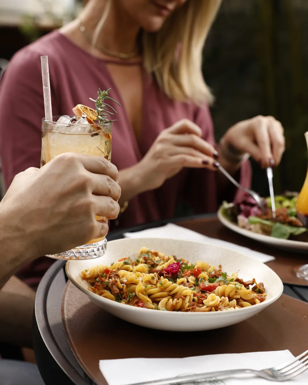 Mesa de restaurante com massa, drink e pessoas compartilhando uma refeição.